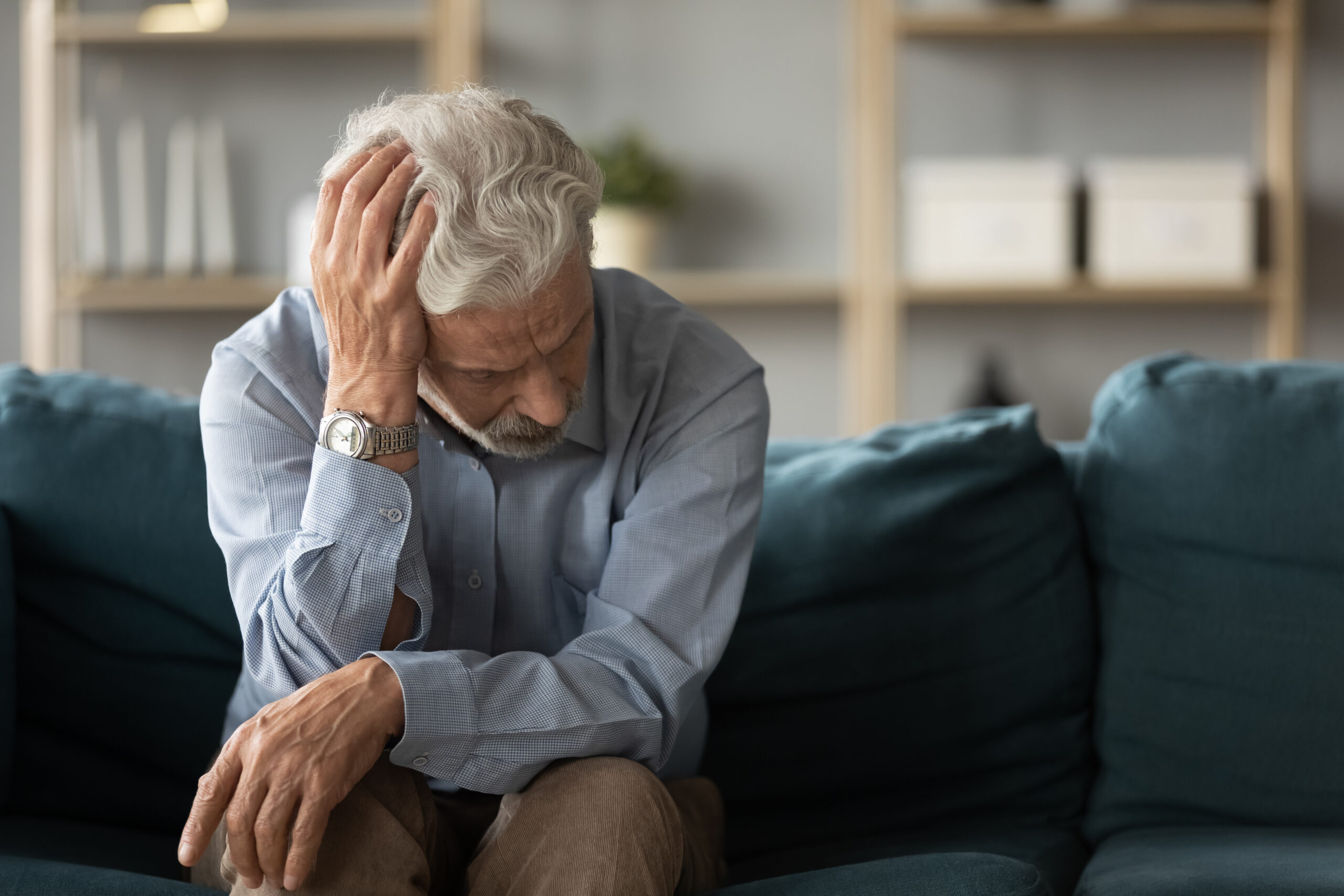 An elderly man sitting alone on a couch with his head bowed and hand on his head, reflecting in silence and solitude.