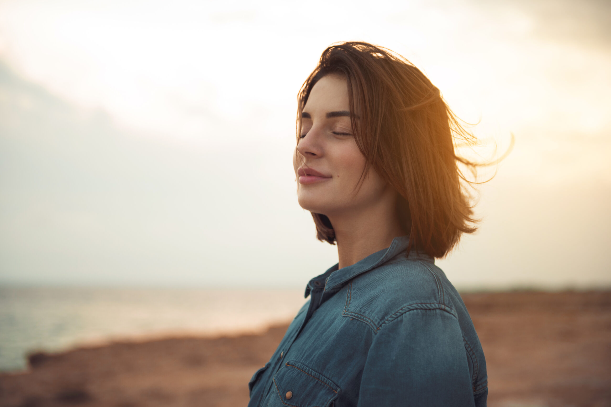 A serene woman stands by the sea at sunset with her eyes closed, symbolizing inner peace and reconnecting with intuition.