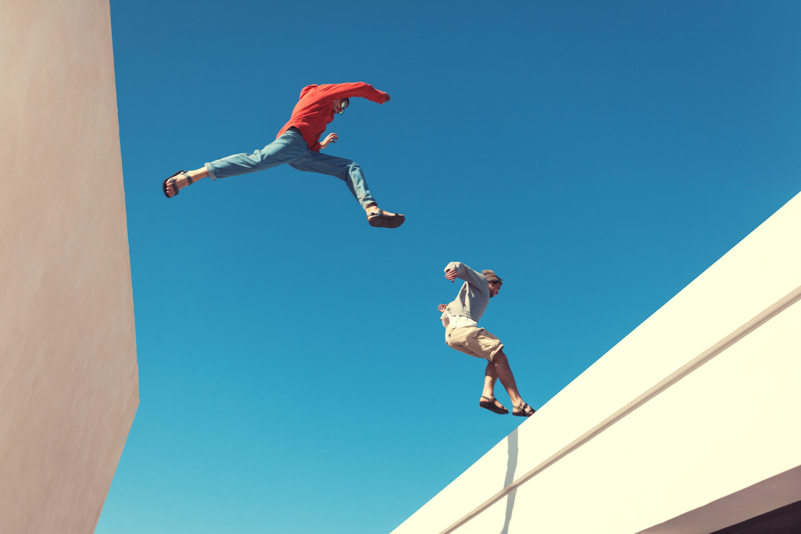 Two people mid-air leaping between rooftops against a clear blue sky, capturing the boldness and trust in gut instinct over calculated thought.