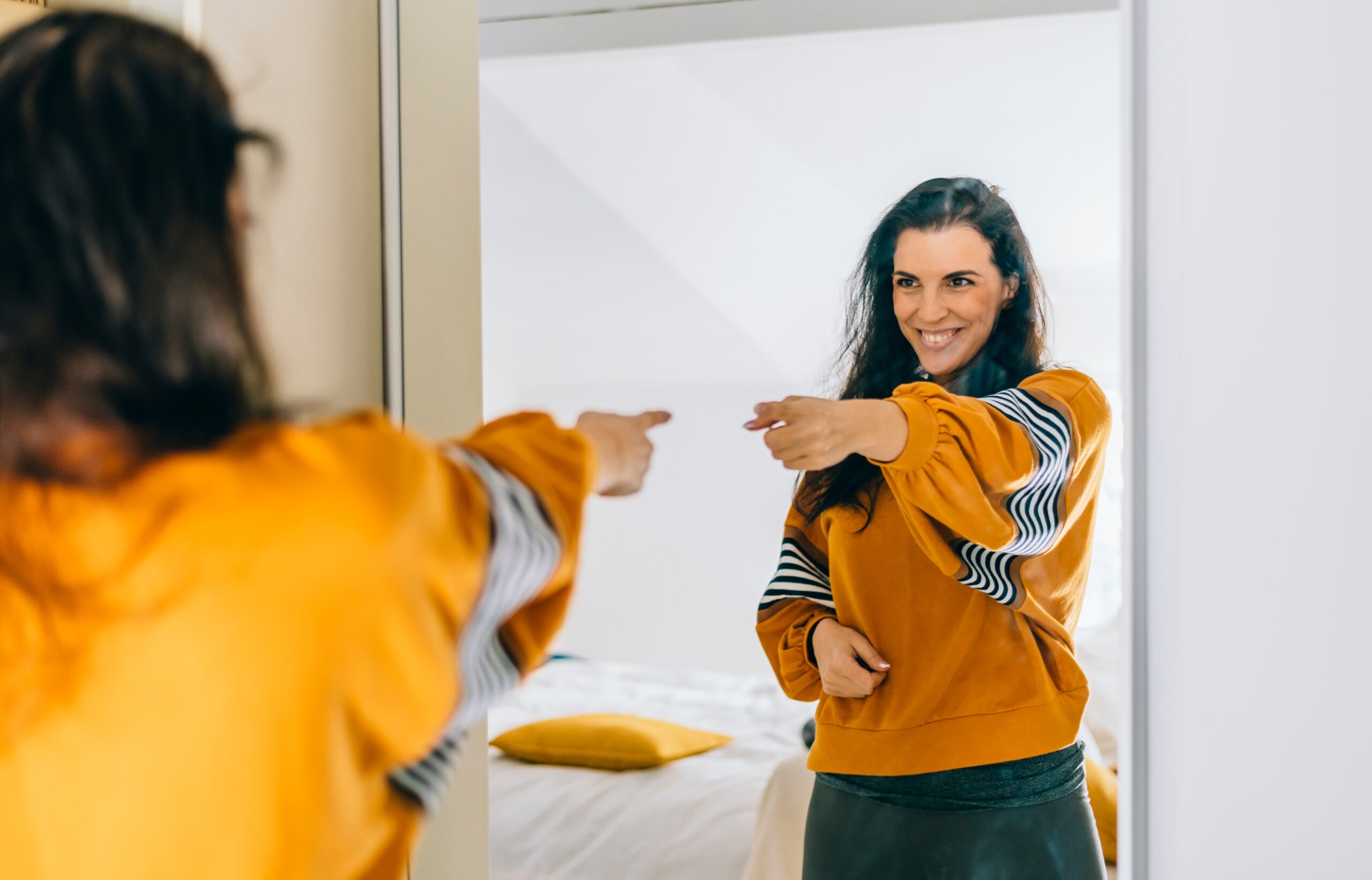 A woman smiling confidently at her reflection in the mirror while pointing at herself, symbolizing the clarity and direction gained through self-reflection.